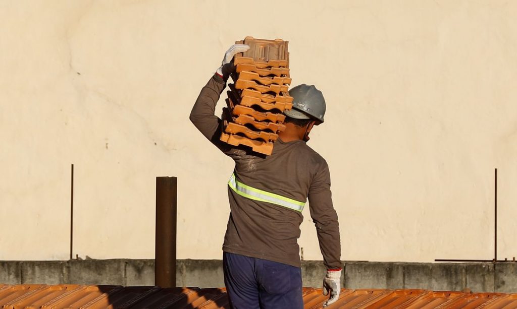 imagem mostra trabalhador carregando telhas nos ombros enquanto usa capacete
