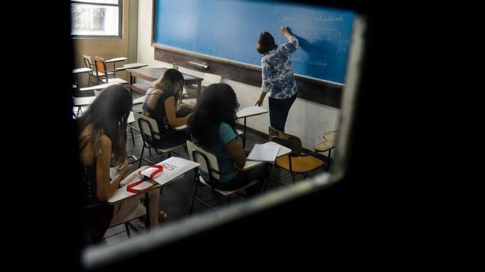 Imagem mostra professora escrevendo em quadro de giz e adolescentes escrevendo em caderno em sala de aula